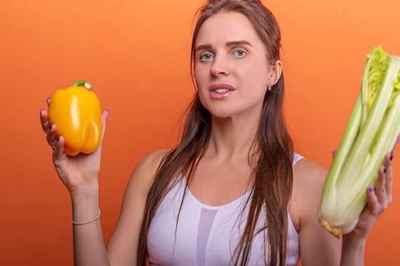 Woman holding yellow bell pepper and celery, showcasing healthy eating.