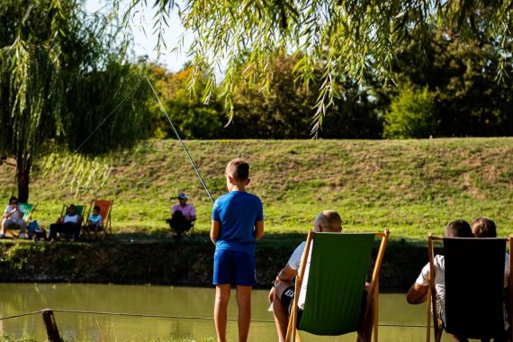 Young boy fishing by a pond on a sunny day, surrounded by trees and relaxed people.